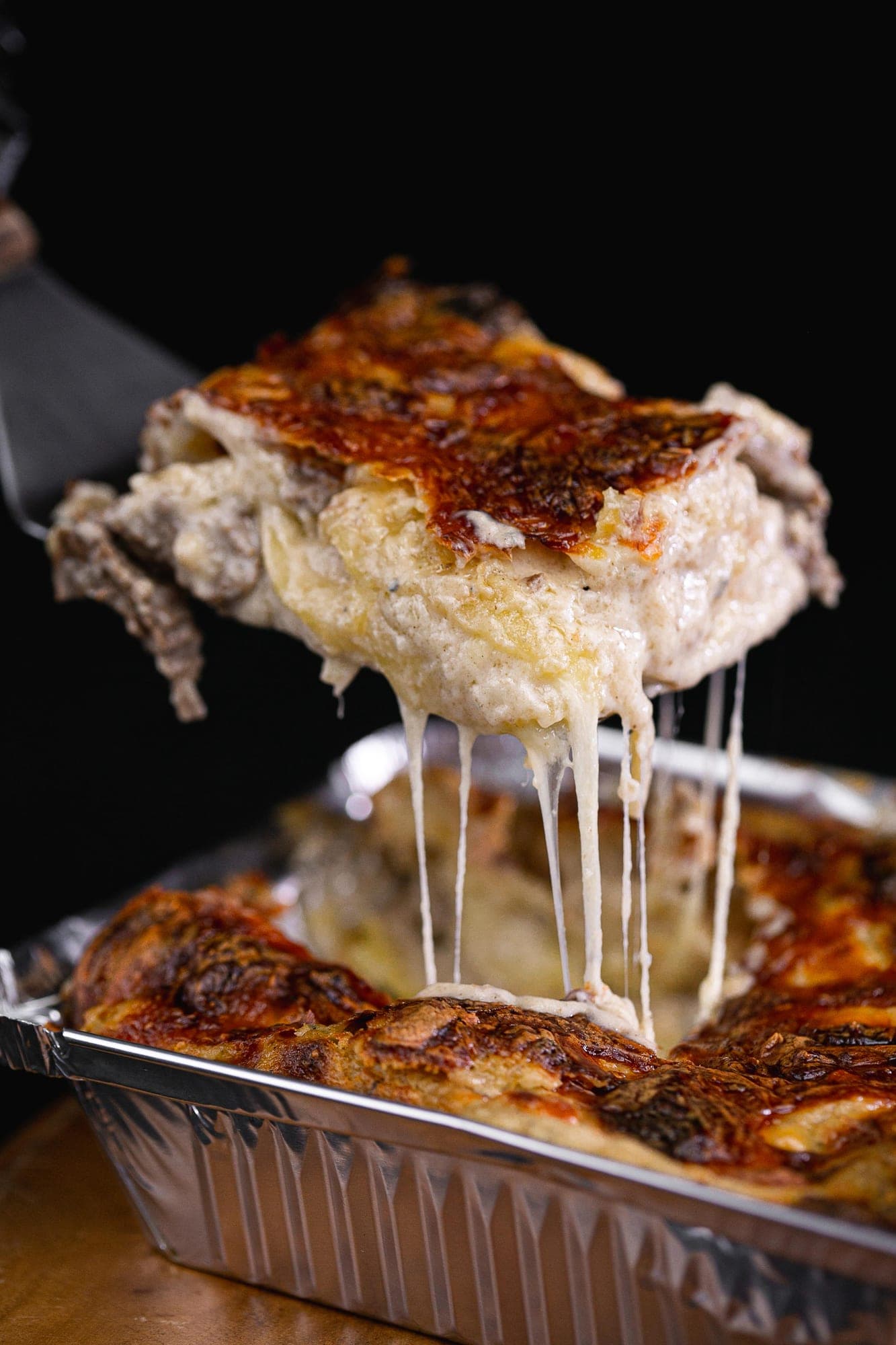 Porção de lasanha de carne gratinada com queijo derretido sendo servida de uma forma de alumínio