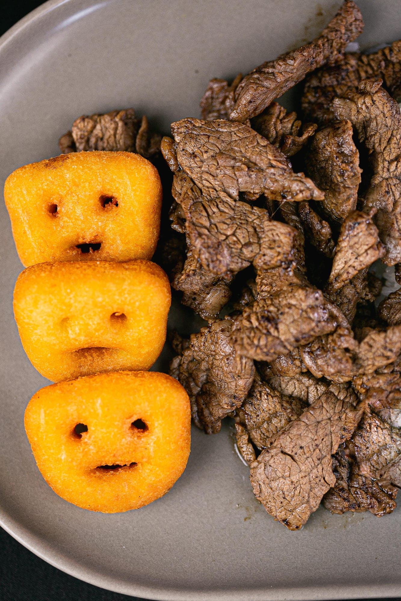 Tiras de carne grelhada ao lado de bolinhos de batata dourados em prato de cerâmica